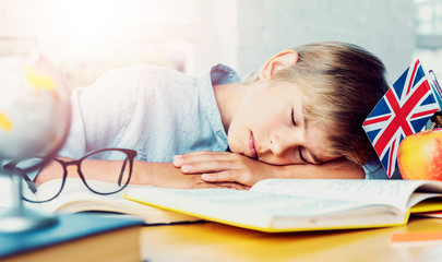 Young tired schoolboy felt asleep in the classroom, table with textbooks and british flag