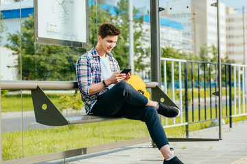 Male student wth smart phone in hands wearing plaid shirt sitting on bench while waiting for...