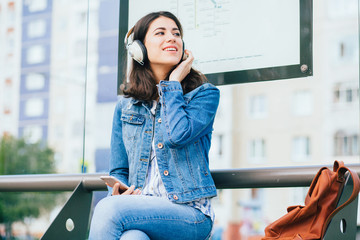 Smiling excited brunette woman wearing blue denim jacket with headphones looking away, relaxing, listening music while waiting at tram stop in a city.