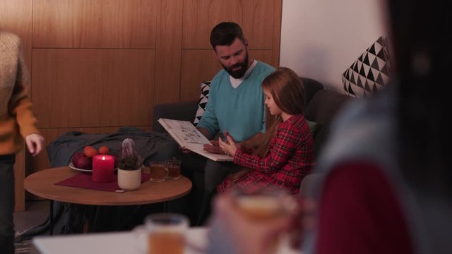 Father with daughter reading children book son make tea on a sofa in evening family home smile woman lamp girl living love male night parenting room sitting young apartment cozy slow motion