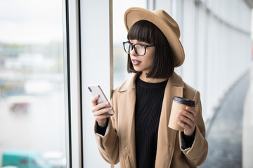 Attractive business woman wear in coat and hat drinking coffee in her office. Pretty young business woman having coffee at her workplace. Female coworker at modern workspace drinking coffee.