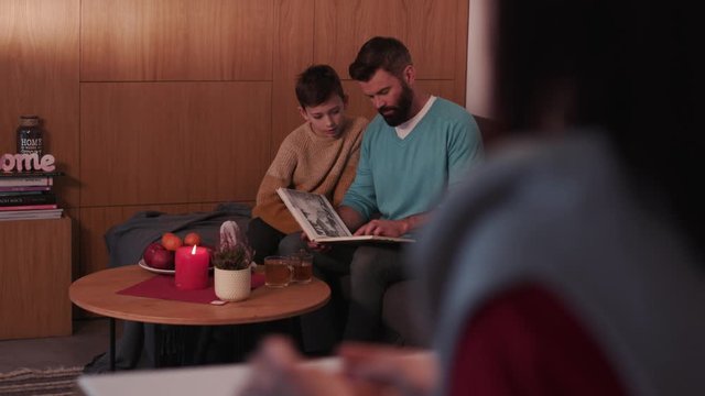 Cute father with son reading children book daughter make tea on a sofa in evening family home smile woman lamp girl living love male night parenting room sitting young apartment cozy slow motion