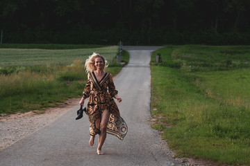 Obraz premium Blonde girl in a patterned dress runs on a country road near the forest