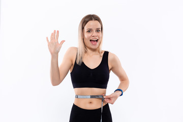 Fototapeta na wymiar Young blond woman in a black sportswear with smart watches is happy checking her waist with tape measure after workout standing over white background.