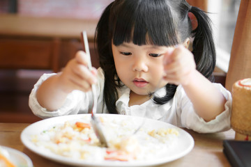 Asian child cute or kid girl enjoy eating shrimp fried rice delicious food with hungry on wood table and white dish for breakfast or lunch and dinner in the cafe restaurant with holding spoon and fork