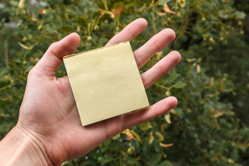 empty yellow sticker pack lies on the palm of a man, green trees in background. small sheet of paper in hand with copy space for text. Hand holding sticky note.