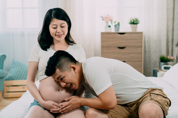 happy handsome asian chinese man listening to beautiful pregnant wife tummy. unborn baby gift for dad on fathers day. charming expectant mom looking at her joyful husband with bare belly sit on bed.