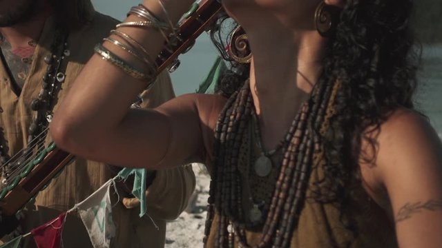 Etnic girl dancing with hands next to the man playing a string on the beach against warm sun