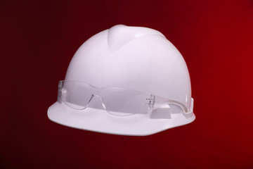 Fototapeta premium White generic protective construction helmet with a plastic transparent safety glasses on the cap against a red studio background. Floating workers hat and spectacles.