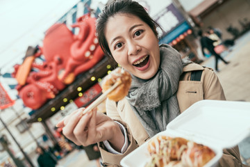 happy young chinese girl holding chopsticks and takeaway box of takoyaki. asian woman tourist face camera smiling and showing tasty octopus balls. japanese food vendor in background with seafood sign
