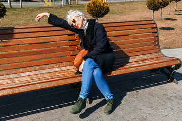 Tired white-haired middle-aged woman resting in a park on a bench after a long day of work. rich Caucasian lady Wearing stylish spring coat and sunglasses. woman fell asleep on a bench in the park.