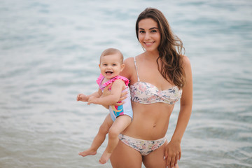 Mother hold cute baby girl on hands on the beach in the sea. Mom and daughter in swimsuit