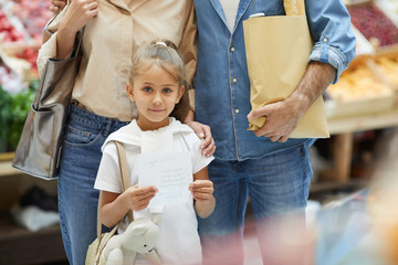 Portrait of cute girl holding shopping list while posing with parents at farmers market, copy space