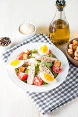 Fresh salad made of tomato, ruccola, chicken breast, eggs, arugula, crackers and spices. Caesar salad in a white, transparent bowl on wooden background
