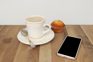 Mock up image of white mobile phone with empty black screen, cup of coffee and cupcake on wooden table