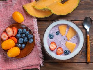 Top view of blueberry smoothie in a coconut bowl with strawberries and melon and kumquat on a red towel with a teaspoon