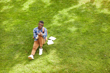 Fototapeta premium Wide angle portrait of African-American student sitting on green grass in campus and doing homework outdoors, copy space