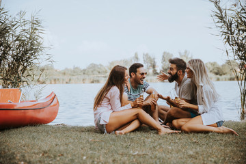 Obraz premium Group of friends with cider bottles sitting by the boat near the beautiful lake and having fun