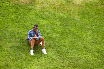 Fototapeta na wymiar Wide angle portrait of young African-American man sitting on green grass in campus and doing homework outdoors, copy space