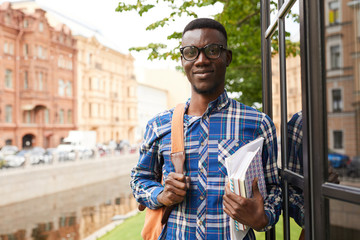 Waist up portrait of smiling African-American student posing outdoors in college campus and looking at camera, copy space