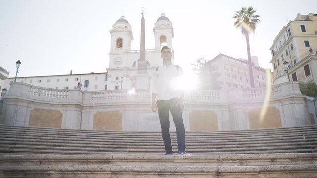 Handsome tall young tourist with black rucksack staying on the stairs in Rome. Amazing ancient background with sunrise behind the man. Morning italian atmosphere insiping young teenager for reaching