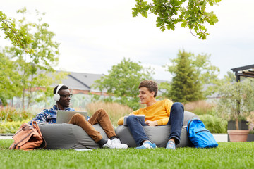 Full length portrait of two international students chilling on bean bags outdoors in college campus, copy space