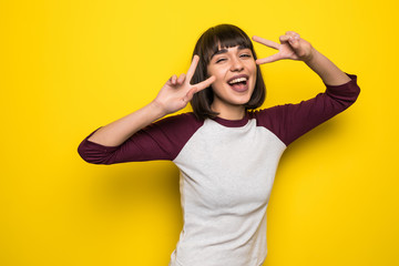 Happy pretty girl posing at camera showing peace sign isolated on yellow background