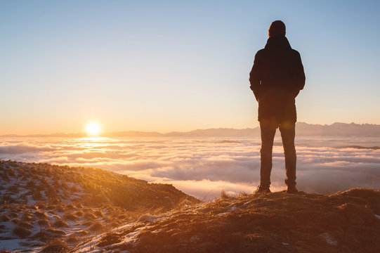 View From the back. A lonely standing man high in the mountains looks at the setting sun and the sunset horizon with a valley filled with clouds. The concept of tourism travel and male loneliness