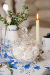 Glass vase with dessert marshmallow is on the table, covered with a tablecloth with a pattern Gzhel. Beside the candle burns. Decor serving a festive dinner or lunch