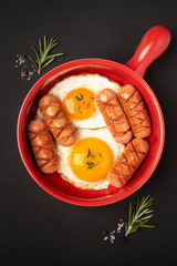 Red pan with fried eggs and sausages on a black background , close-up. English breakfast.