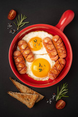 Red pan with fried eggs and sausages on a black background , close-up. English breakfast.
