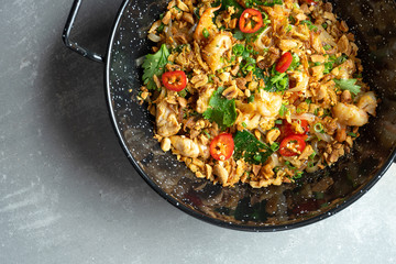top view on Pad thai, or phad thai stir-fried rice noodle with vegetables and chicken, hot chili pepper and parsley in black bowl on grey stone background. Street pan-asian food with copy space
