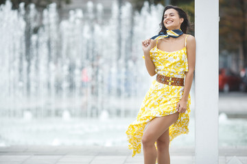 Attractive young woman near the fountain. Beautiful female outdoors. Lady in yellow dress portrait.