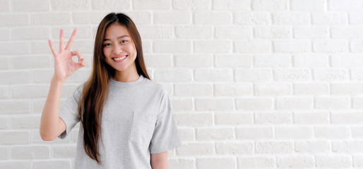 Young asian woman showing ok hand sign and smiling while standing over white brick wall background with copy space, Happy asian girl showing ok hand, Positive people gesture