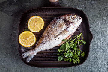 Raw dorado fish with spices, lemon and parsley in black grill pan on black concrete background. Top view