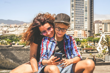 Fototapeta premium Mother and son watching smartphone together sitting outside in the city. Woman and trendy young boy playing with cell phone. Family video call during summer holiday New technology social media concept