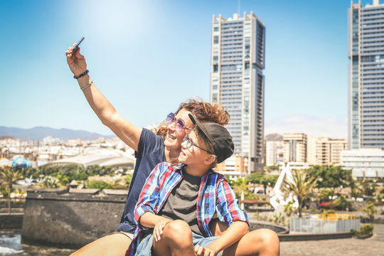 Mother and son taking selfie with smartphone sitting outside in the city. Woman and trendy young boy playing with cell phone Family video call during summer holiday New technology social media concept