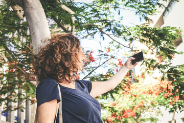 Young woman taking selfie with smartphone outside in the city. Trendy girl playing making new trend video story with cell phone under flowering trees. New technology and modern communication concept