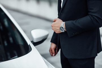 Close up of a man with a watch in his hand in a business suit near a premium white car.