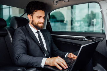 Handsome, bearded, smiling manager working on his laptop on the backseat of the car.