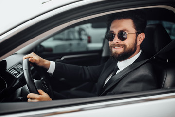 Handsome young smiling bearded driver in full suit with fastening seat belt driving a new white car.