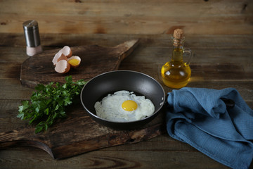 frying pan with frying egg with yolk on a wooden cutting board with ingredients for the preparation of fried eggs: raw eggs, olive oil, salt, parsley. With copy space for text or image.