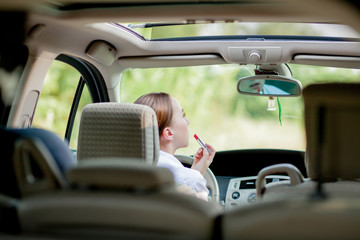 Concept of danger driving. Young woman driver red haired teenage girl painting her lips doing...