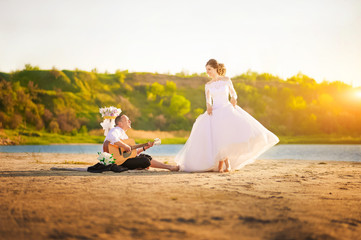 Fototapeta na wymiar Romantic wedding couple at sunset on the shore with a guitar. The bride and groom near the shore on a sunset beach in summer.