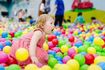 Portrait little cute baby girl princess infant 1-2 year standing and play with balloons, colorful balls in playground, ball pit, dry pool for birthday party. Celebration first year concept.