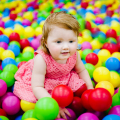 Fototapeta na wymiar Happy laughing girl playing with toys, colorful balls in playground, ball pit, dry pool. Little cute child having fun in ball pit on birthday party in kids amusement park and play center indoor.