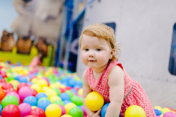 Fototapeta na wymiar Portrait little cute baby girl princess infant 1-2 year standing and play with balloons, colorful balls in playground, ball pit, dry pool for birthday party. Celebration first year concept.