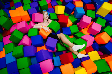Fototapeta na wymiar A young woman or girl, mother playing and jumping in soft cubes in the dry pool of the game children's room for birthday. entertainment centre. indoor playground in foam rubber pit in trampoline.
