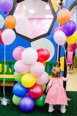 Fototapeta na wymiar Portrait little cute baby girl princess infant 1-2 year standing back and playing in the gaming room, with balloons, colorful balls in playground in birthday party. Celebration first year concept.