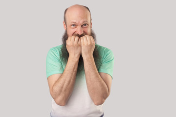 Portrait of nervous middle aged bald man with long beard in light green t-shirt standing, bitting...